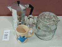 Photo showing the aluminum coffee maker, Pyrex glass coffee pot, and Cookie Monster themed mug on a white surface against a red wall.