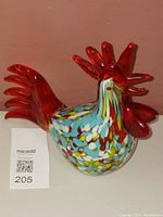 Front and side view photo of the glass rooster figurine on a solid surface showing its vibrant red comb and tail with speckled body.