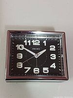 Front view of Seiko square desktop clock with black dial, white numbers, and silver frame.
