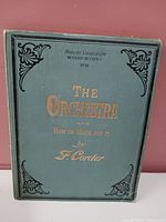 Front cover of the cloth-bound book 'The Orchestra and How to Write for It' by F. Corder showing green cover with gold embossed title and decorative corners.