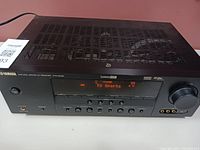 Front and top angled view of Yamaha HTR-6025 AV receiver showing control buttons, digital display, and ventilation on top. Unit powered on displaying 'TV Sports'.