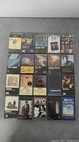 20 various cassette tapes from classic rock and pop artists arranged in 4 rows on a gray surface. Visible titles include The Cars, George Carlin, Genesis, Earth, and Peter Gabriel.