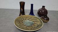 Five ceramic and pottery items arranged on a gray surface showing the raised plate and four vases with varied glazes and shapes.