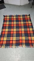 Full view of the large wool blanket laid flat on floor showing the plaid pattern in orange, yellow, and dark blue with fringe edges.