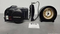 Front and side view of Olympus camera with attached handgrip and lens cover next to Pottery Barn black desk clock with gold-tone rim.