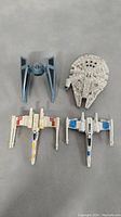 Top view of all four Star Wars fighter models laid out on gray surface: TIE Fighter, Millennium Falcon, one red X-Wing, one blue X-Wing.