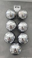 Top-down view of all 7 shiny metal bocce balls arranged in pairs and singly, showing reflective surface and incised line patterns.