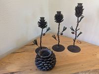 Four metal candle holders with pine cone themes; three tall with pine cones on stems decorated with leaves and red berries, one round pine cone-shaped holder on a wood table near cream wall.