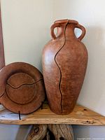 Photo showing large two-handled brown pottery vase with wavy vertical line design next to a matching round plate with similar wavy line pattern on a wooden shelf.