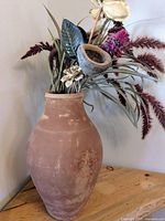 Photo of large terracotta pottery vase with artificial floral arrangement on wooden surface against beige wall.