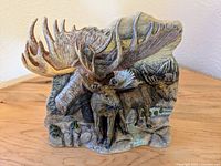 Front view of the pottery sculpture showing two moose in a natural mountainous forest setting with detailed textured antlers, fur, trees, and rocks.