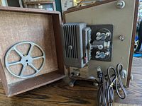 Photo of vintage film projector alongside its original carrying case, showing front and top views of the projector and interior of the case.