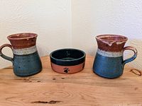 Full view of two ceramic mugs and one ceramic bowl arranged on wooden surface against a white wall.