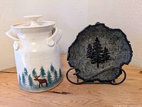 Two-piece pottery decoration set: white ceramic canister with moose and tree painting plus leaf-shaped stoneware plate with tree design on metal stand.