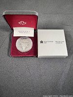 The uncirculated Regina coin displayed inside a protective case with a red velvet interior, accompanied by a small inspection slip inside the case and a white Royal Canadian Mint branded box beside it.