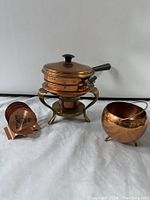 Three copper items placed on white cloth: copper bowl with handle, copper chafing dish on brass stand with lid and black handle, and copper coaster holder with coasters.