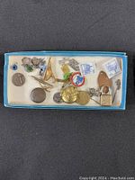 Square photo showing an array of various small items in a blue box on a dark surface, including jewelry pieces, coins, charms, a small padlock, and two stamps or tags.