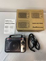 Photo showing the Audiocrazy portable radio with power cable, original box, and instruction manual.