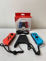 Photo showing the Joy Pad controllers in orange and blue colors, the black central grip, charging cable, and packaging box.