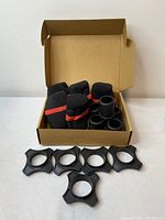 Open cardboard box containing five black microphone foam covers with a red stripe and five black plastic mounting adapters laid out in front.