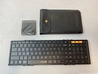Top view of the unfolded ProtoArc folding keyboard, black with orange key highlights, alongside a black carrying case and a screen protector still in packaging.