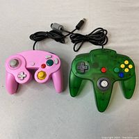 Top view of pink GameCube and green translucent Nintendo 64 wired controllers showing buttons and joysticks.