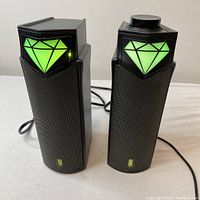 Front view of the two black USB computer speakers showing vibrant green diamond-shaped LED lights on each speaker, and a volume control knob on the right speaker.