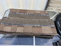 Stack of 25 tan asphalt shingles showing granulated texture and adhesive strips on the back, placed on a surface near a container.