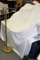 Full view of the brass floor lamp standing next to a covered couch, showing the polished circular base and adjustable swing-arm but no lampshade attached.