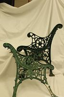 Two vintage green cast iron bench ends with intricate Victorian scroll and floral design shown side-by-side against beige backdrop.