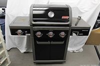Front view of Coleman Revolution BBQ grill showing four main burners, side burner, and control knobs, with closed hood and side shelves.