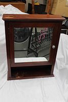 Front view of wooden wall cabinet showing mirrored door with silver knob, open bottom shelf, and signs of wear on the top surface.