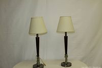 Pair of classic table lamps with tapered dark wooden bases on polished metal round stands and cream fabric drum-shaped shades placed on neutral cloth background.
