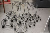 Eight black metal plant stands arranged on floor, showing variety of sizes and styles including taller stands with wire-frame legs and shorter round grid bases with wheels.