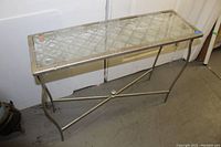 Full view of silver metal console table with lattice glass insert and cross brace base on a concrete floor.