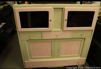 Photo of vintage two-tone wood cabinet painted pale green and soft pink, showing front view with glass upper doors, drawers, and lower cabinet doors.