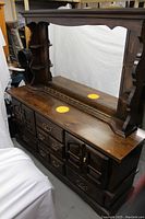 Front angle view of the wooden dresser showing its dark finish, multiple drawers, brass handles, large framed mirror, and built-in shelving on sides of the mirror.