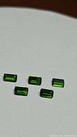 Photo of five small green rectangular baguette cut stones arranged on a white background highlighting the electric green color and clarity of the chrome diopside gemstones.