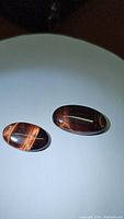 Two oval cabochon natural red tiger's eye gemstones shown on white background with visible chatoyant bands and polished surfaces.