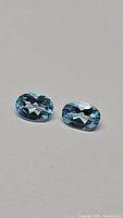 Two natural oval cut sky blue topaz stones placed on a gray background showing facets and matched color pair.