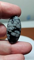 Oval polished cabochon Snowflake Obsidian gemstone held between fingers showing snowflake crystal patterns.