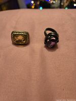 Photo of two heavy rings showing a rectangular yellow gemstone on a gold-tone band and a round purple gemstone on a silver-tone band.