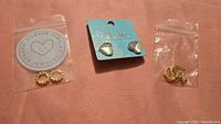 Three pairs of earrings arranged on a pink surface: gold-tone textured hoops, silver acorn studs on a card, gold-tone hoops with moon and star charms in plastic bag.