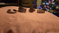 Four metal bracelets laid out on a pink fabric surface in front of a holiday tree.