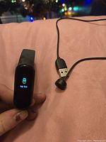 Amazfit smart band with screen displaying 'Pair First' and USB charging cable with magnetic charger end placed on a pink surface.