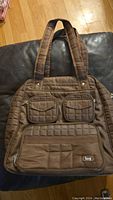 Front view of a brown quilted nylon Lug bag with dual top handles, two small zippered pockets at the upper front, a larger zippered pocket with logo below, and two snap pockets at the bottom front.