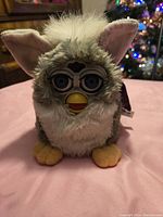 Front view of original 1998 Furby toy showing green and white fur, blue eyes, yellow beak, and feet on pink surface with Christmas decorations in background.