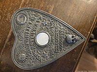 Top view of the heart-shaped metal box featuring Ouija board letters, numbers, a skull, sun, and moon engravings.