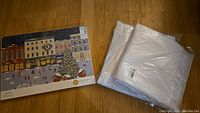 Photo showing boxed set of four Simons placemats with winter holiday village scene and two white packaged tablecloths placed on wooden floor.
