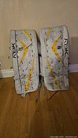 Photo of a pair of white and yellow Reebok adult goalie pads standing upright on a wood floor.